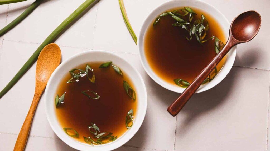 Two bowls of clear broth garnished with chopped green onions, each with a wooden spoon beside or in the bowl, placed on a tiled surface.