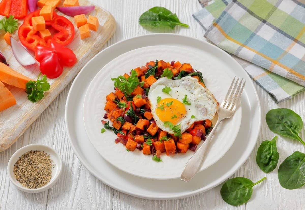 Plate of vibrant spinach sweet potato hash with a sunny-side-up egg and greens, surrounded by fresh vegetables and spices on a wooden table. Cozy and inviting.