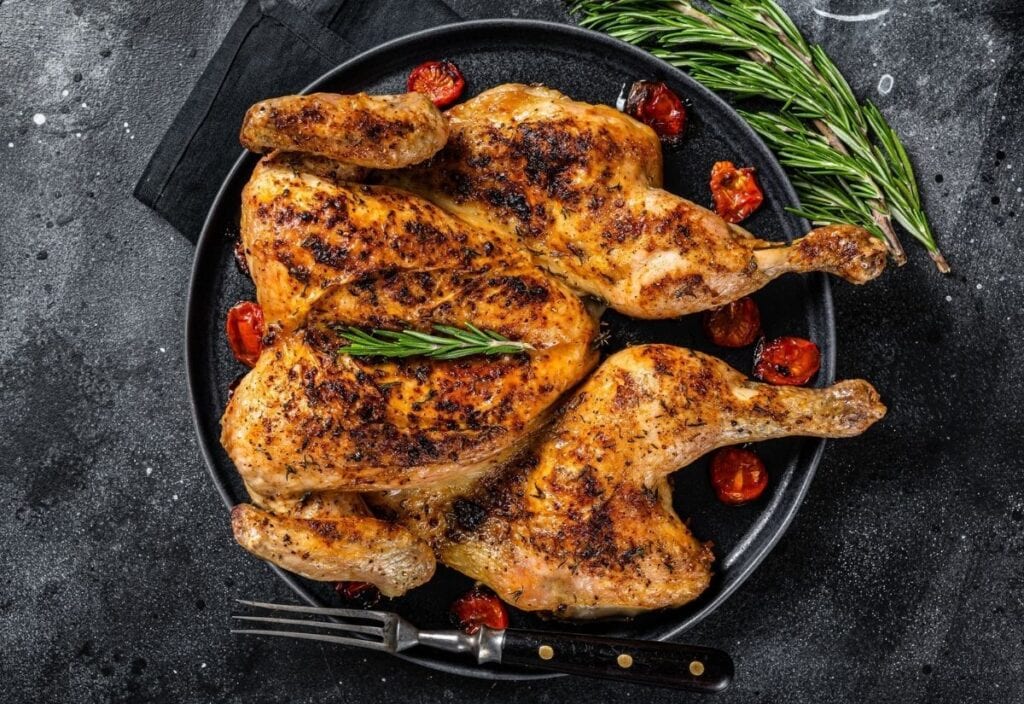 A roasted spatchcock chicken served on a black plate with cherry tomatoes, garnished with rosemary, next to a knife and sprigs of fresh rosemary.