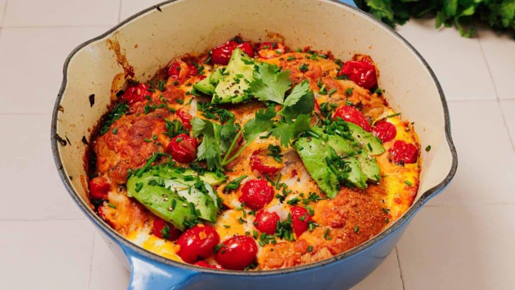 A baked casserole in a blue pot topped with avocado slices, cherry tomatoes, chopped herbs, and sprigs of cilantro.