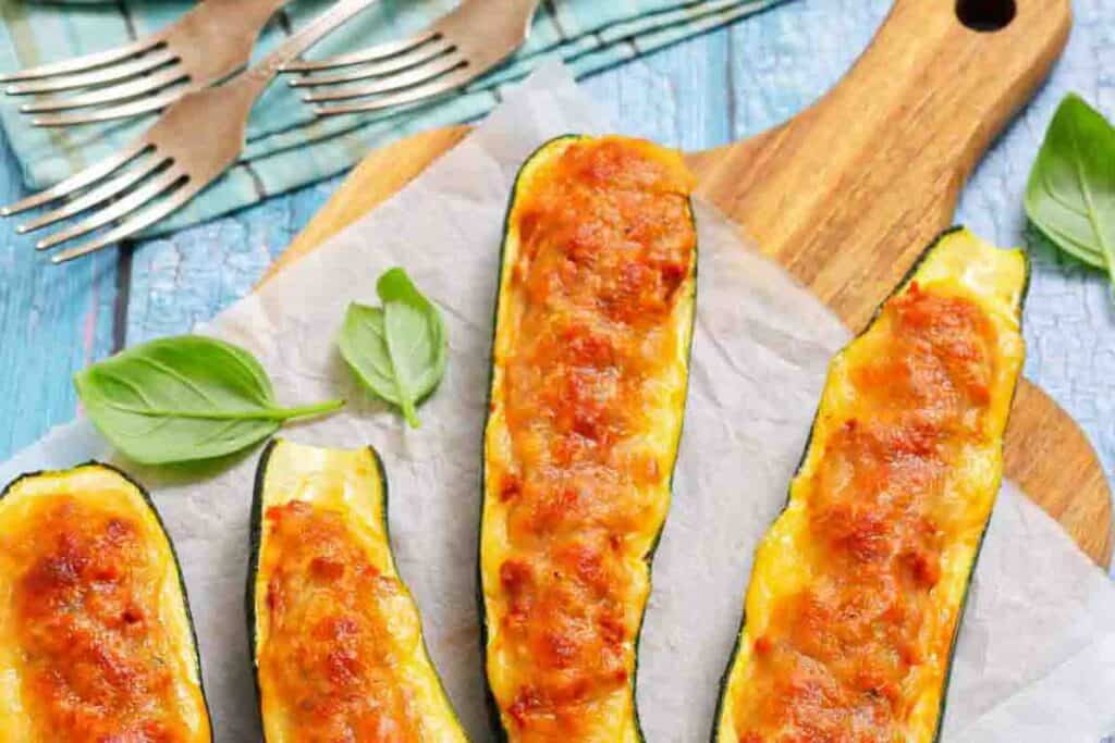 Four baked zucchini boats filled with melted cheese are arranged on a wooden board with basil leaves and forks nearby.