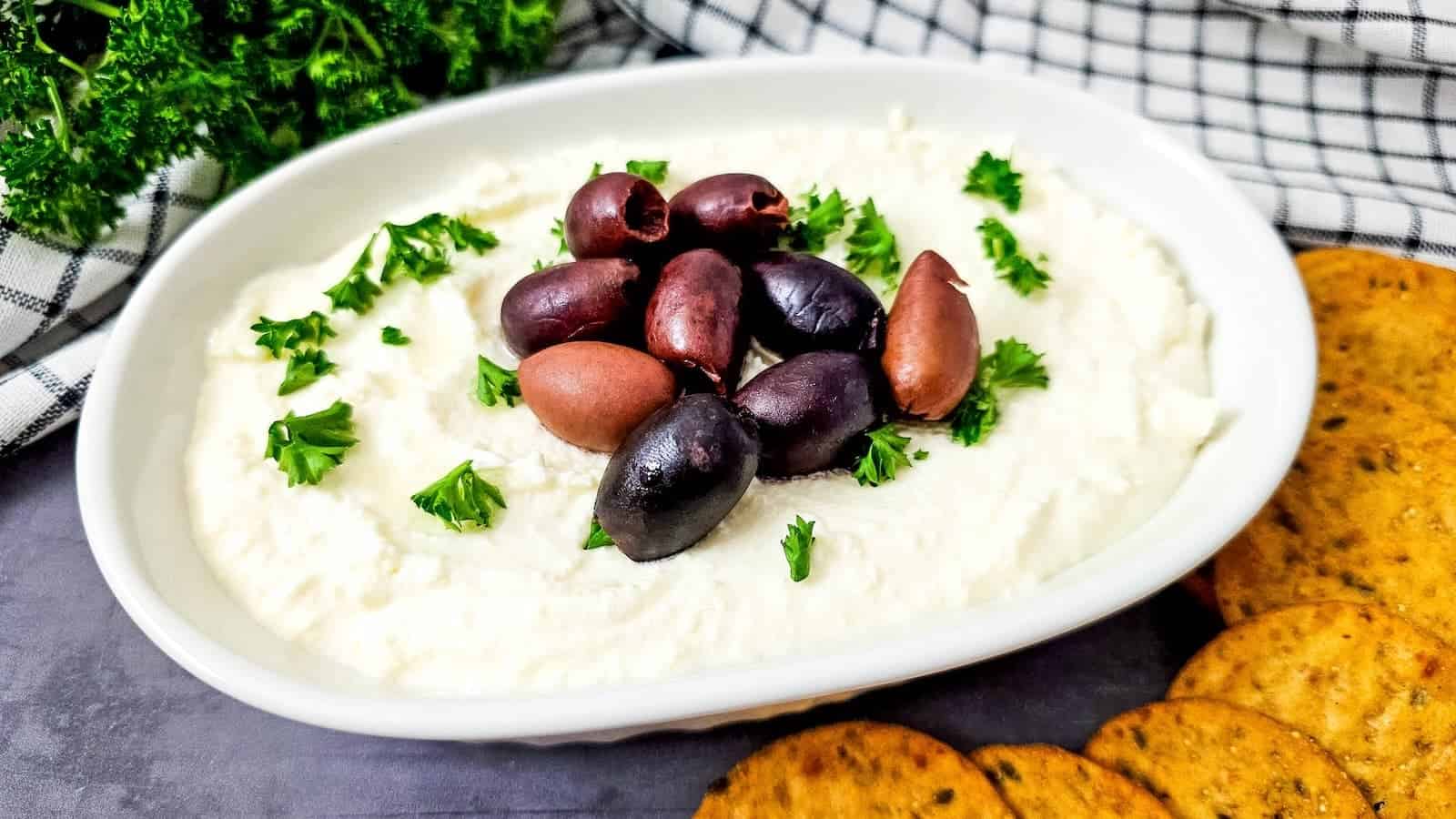 A white bowl of feta dip topped with mixed olives and chopped parsley, placed next to round crackers on a gray surface.