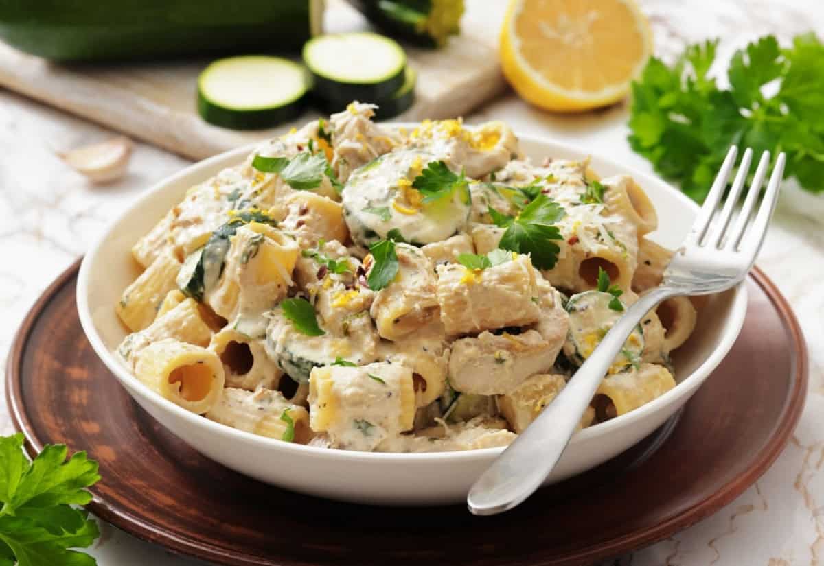 A creamy pasta dish with zucchini, parsley, and lemon zest in a white bowl on a wooden plate. A fork rests on the bowl's rim, creating a fresh, appetizing look.