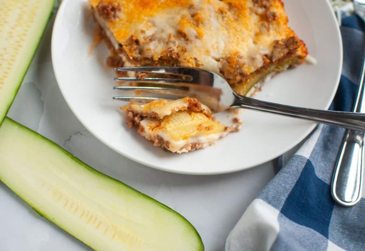 A slice of zucchini lasagna on a white plate with a fork holding a piece, next to fresh zucchini slices and a blue checkered napkin.