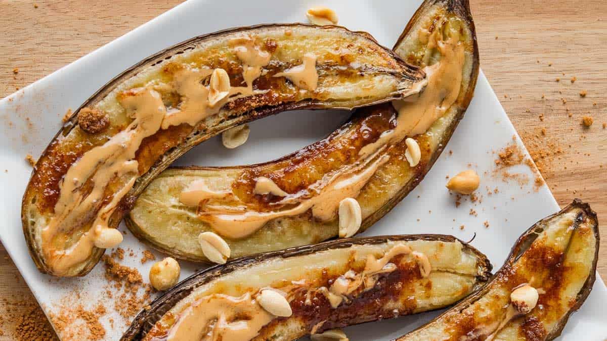 Three halved baked bananas on a white plate, topped with peanut butter drizzle and peanuts, with a sprinkle of cinnamon on the plate.