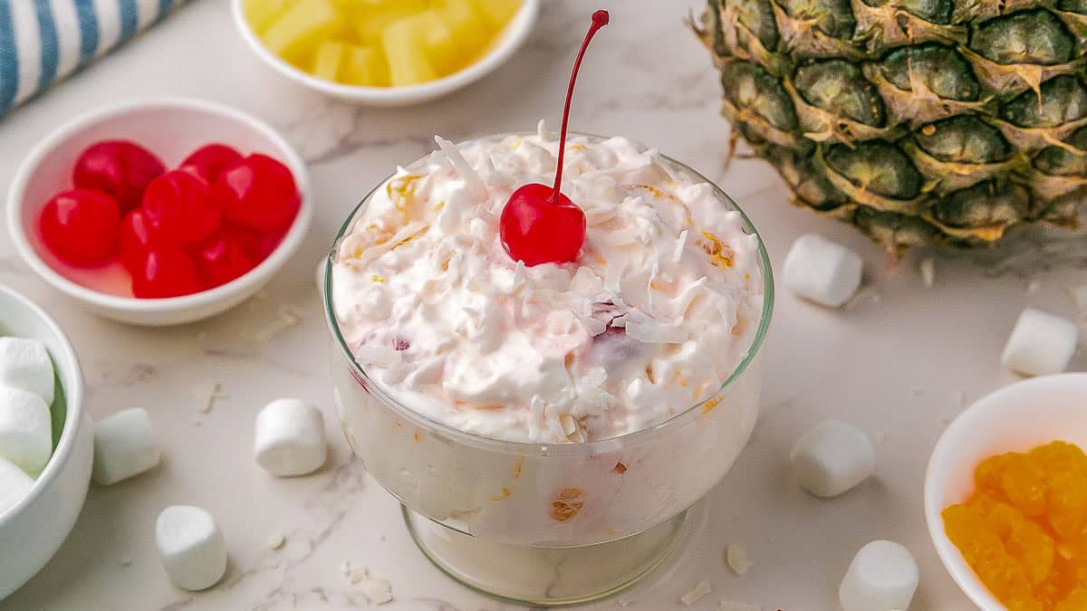 A glass dessert bowl of ambrosia salad topped with a cherry, surrounded by marshmallows, pineapple, cherries, and a whole pineapple on a white surface.
