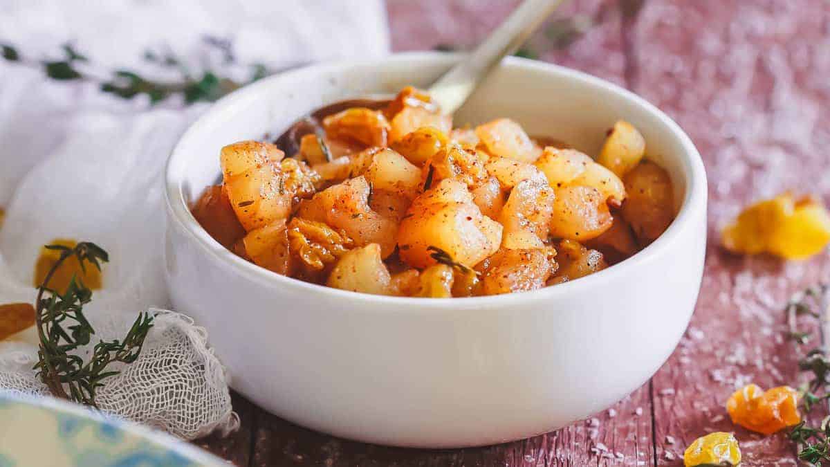 A white bowl filled with spiced, cooked apple pieces, garnished with herbs, with a spoon inside. The bowl is on a rustic wooden surface.