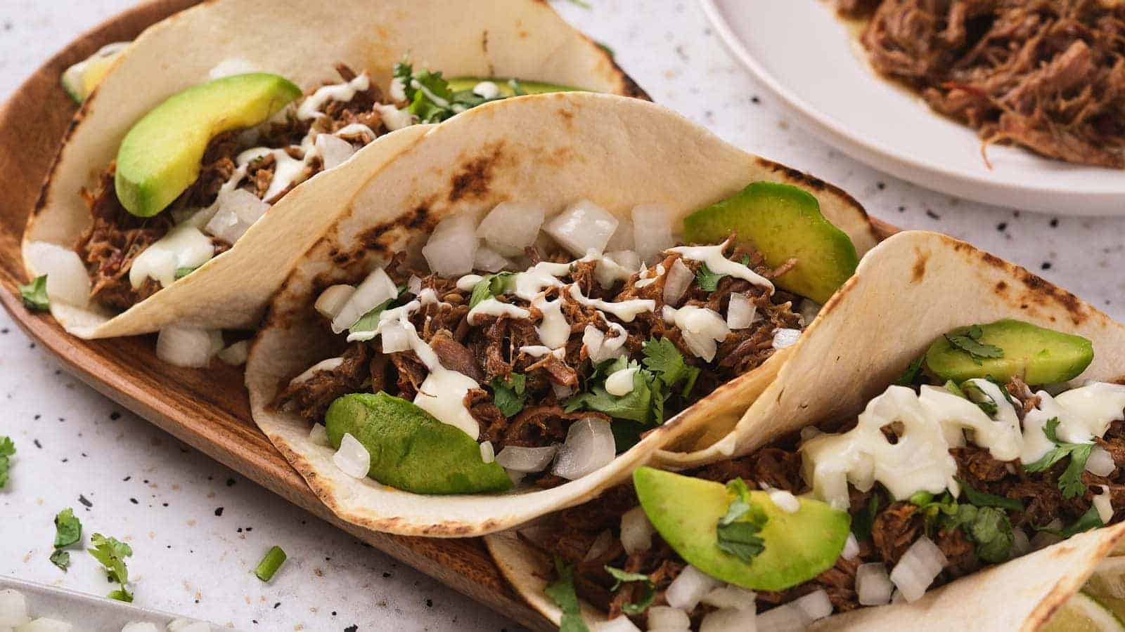 Three barbacoa tacos garnished with avocado, cilantro and white chopped onions next to a plate of shredded beef on the background.