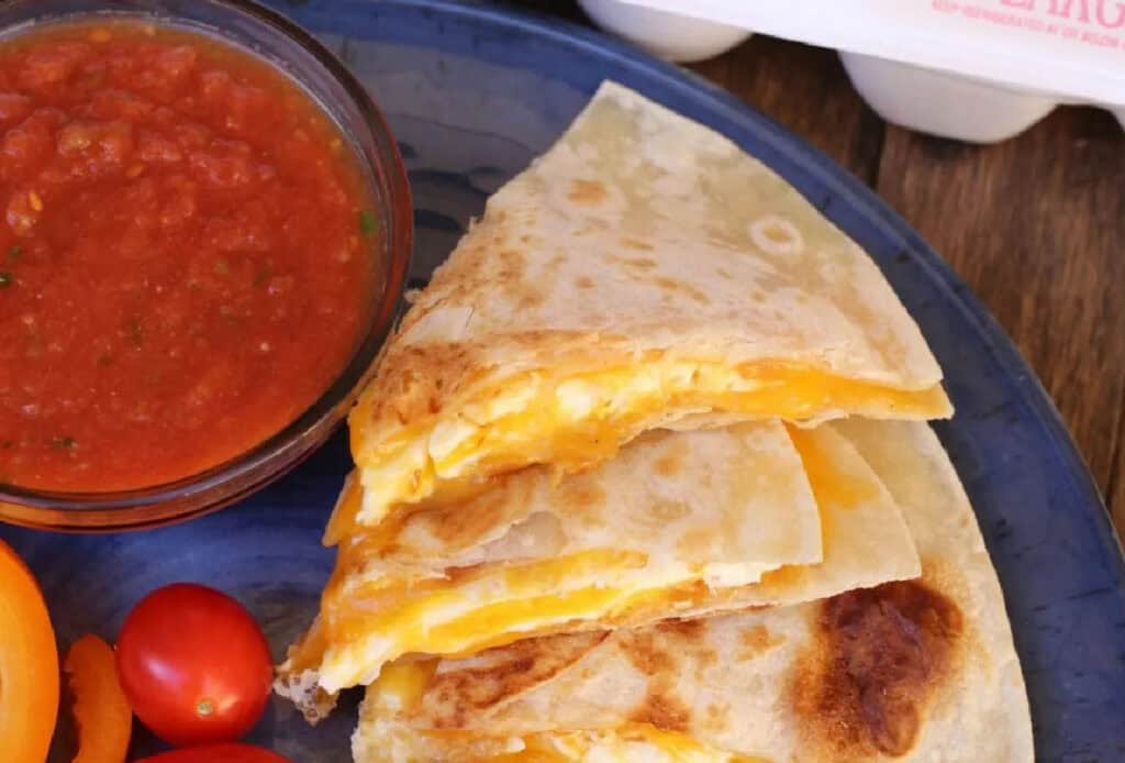 Three cheese quesadilla wedges are stacked on a blue plate next to a bowl of salsa and some cherry tomatoes.