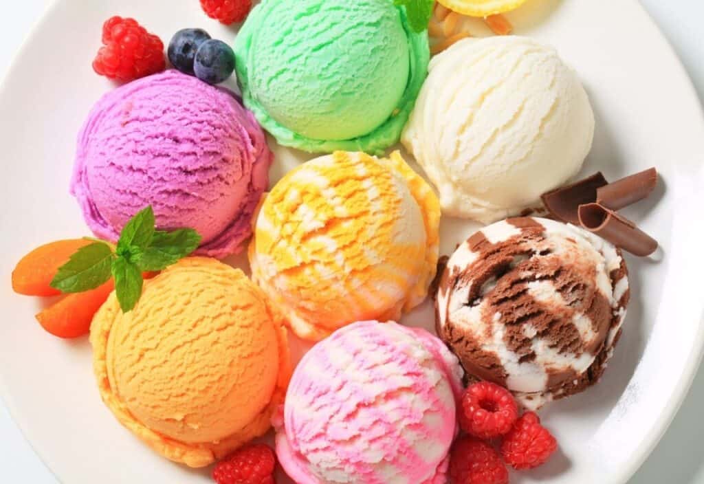 A white plate with six colorful scoops of frozen custard, assorted fresh fruits, and chocolate curls arranged around the scoops.