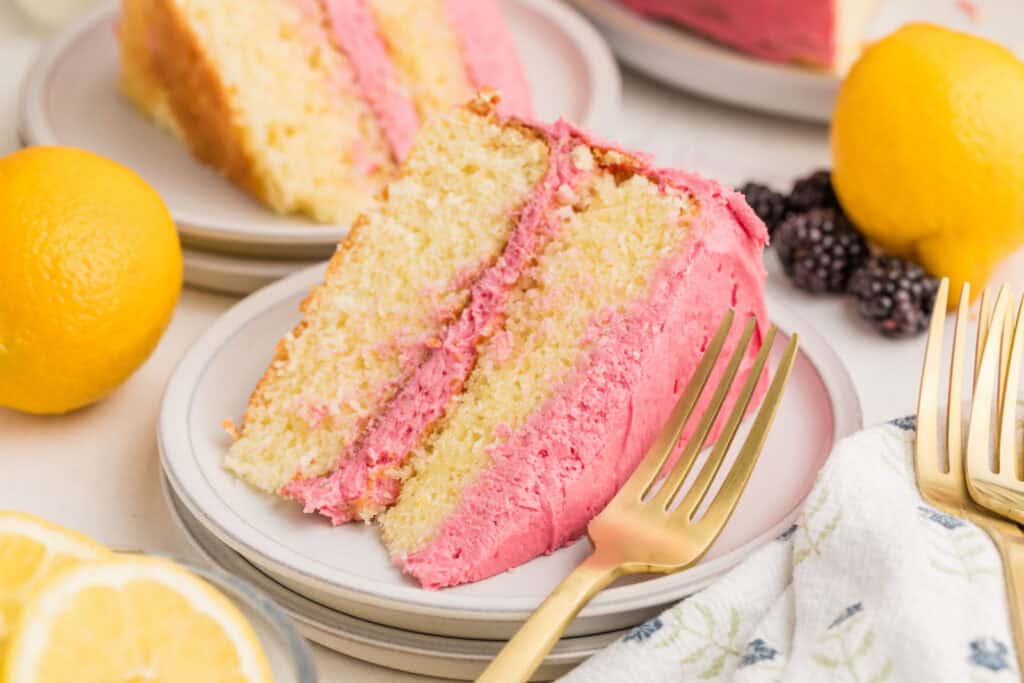 A slice of vanilla cake with pink frosting is served on a plate with a gold fork, surrounded by lemons, blackberries, and another slice of cake in the background.