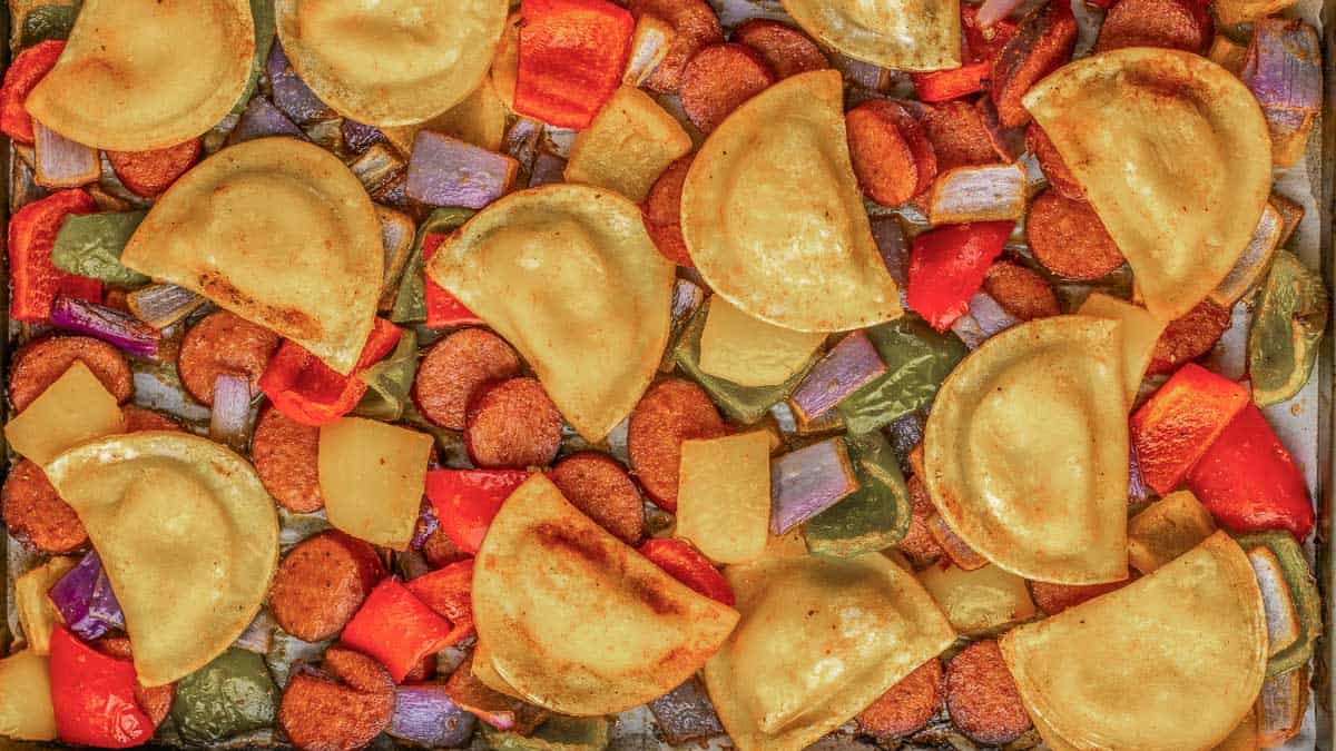 A baking tray filled with cooked pierogi, sliced sausage, red and green bell peppers, onions, and potatoes.
