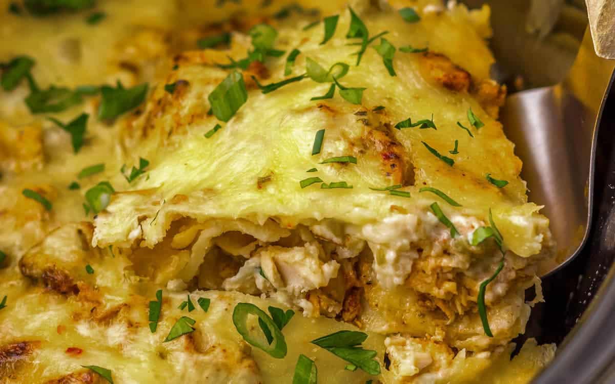 Close-up of a cheesy chicken enchilada casserole topped with melted cheese and chopped fresh herbs, with a serving being lifted from the dish.