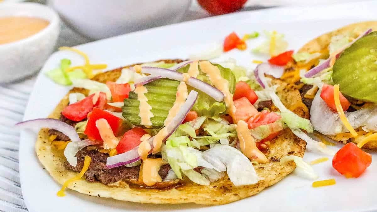 A taco topped with sliced pickles, shredded lettuce, diced tomatoes, red onion, shredded cheese, and a drizzle of sauce on a white plate.