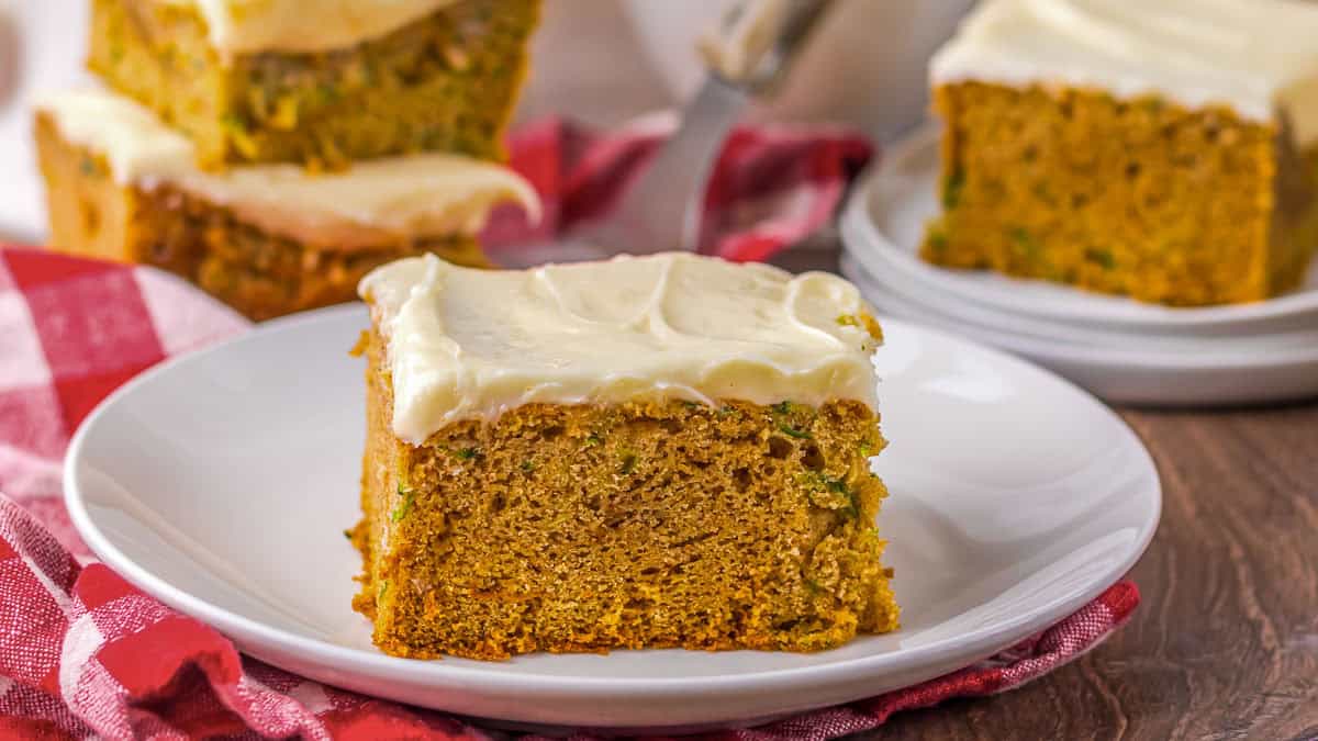 A square piece of frosted cake sits on a white plate, with more pieces of cake and a red checkered cloth in the background.
