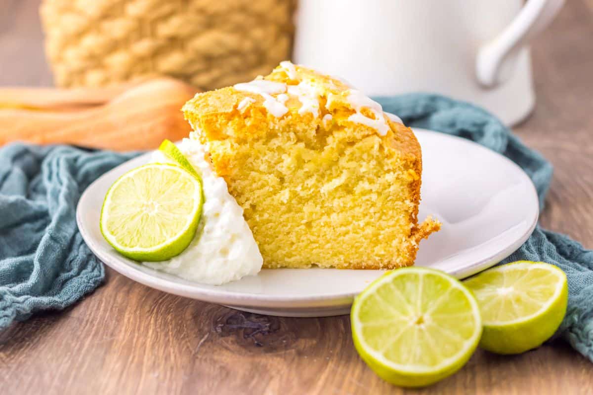 A slice of yellow cake with white icing on a plate, garnished with whipped cream and lime slices, set on a wooden table.
