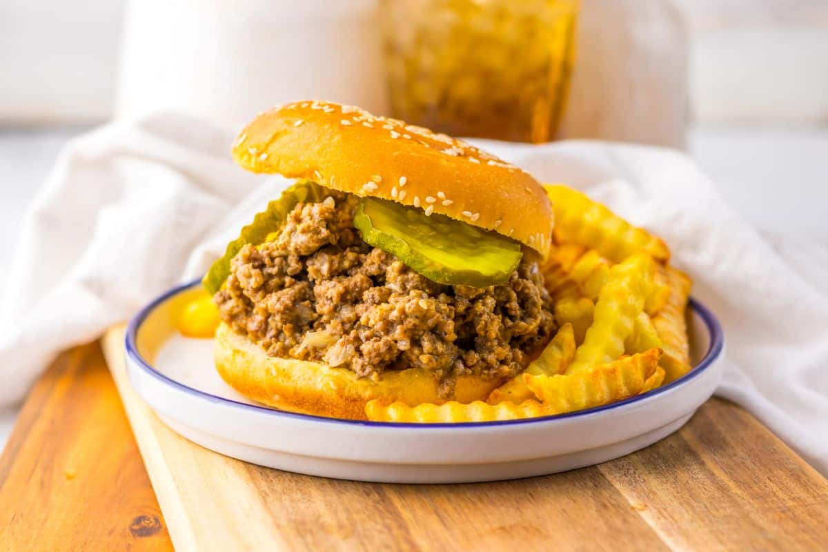 A sloppy joe sandwich with pickles on a sesame bun served with crinkle-cut French fries on a plate.