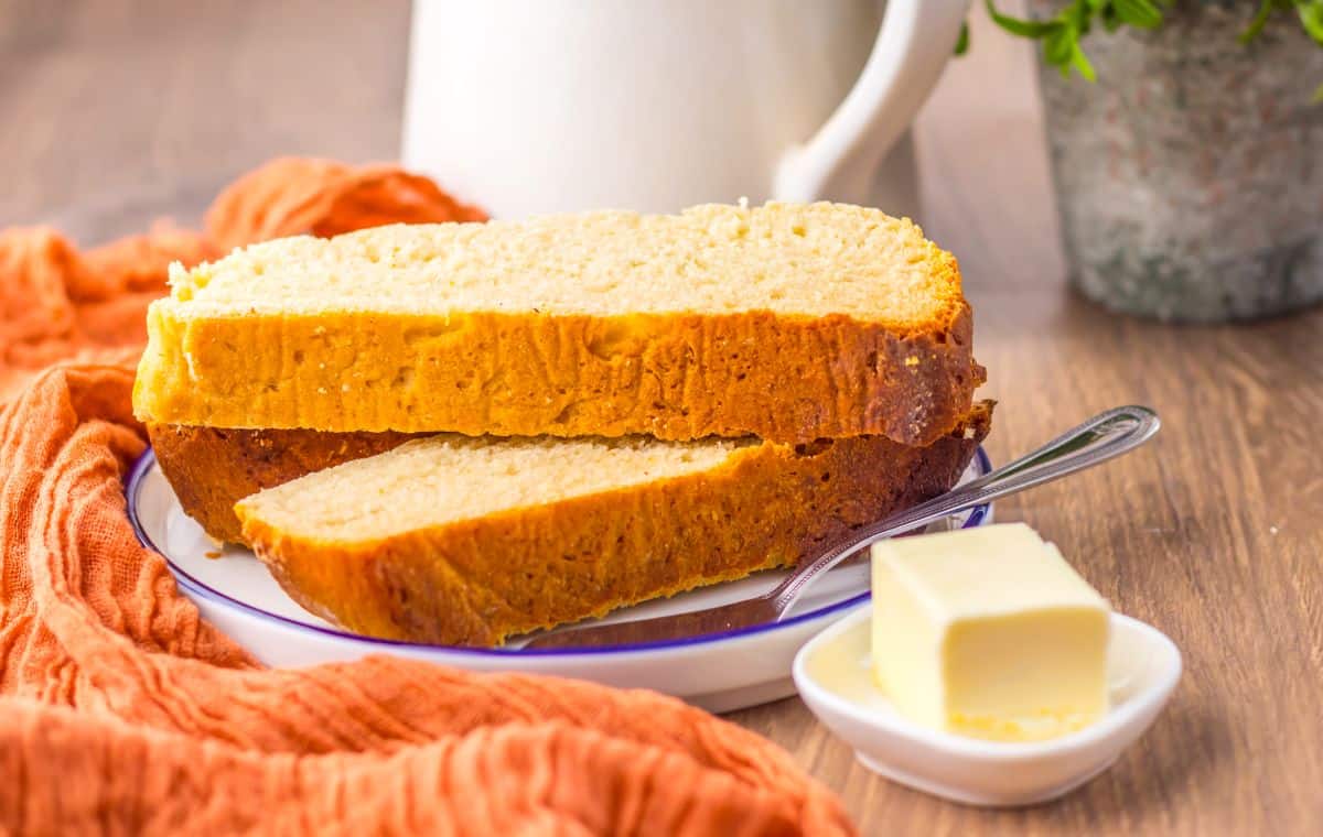 Two slices of bread on a plate with a butter knife, next to a small dish of butter, an orange cloth, and a white pitcher in the background.