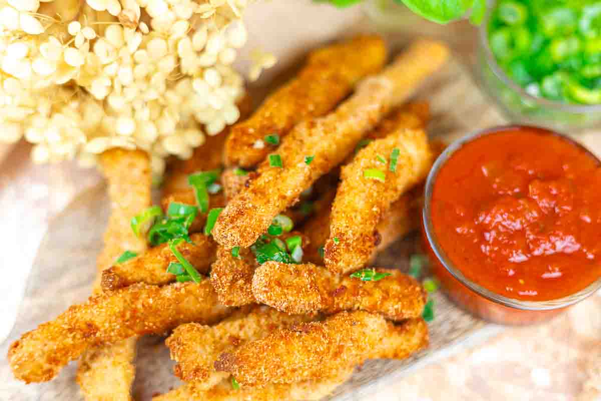 Breaded and fried spicy chicken garnished with chopped green onions, served on a wooden board with a bowl of red dipping sauce.