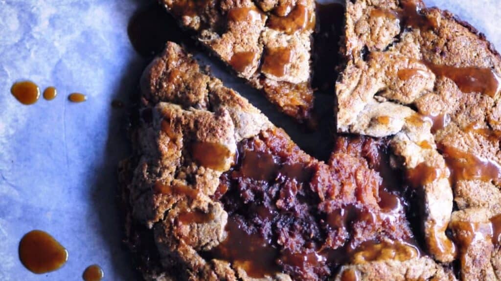 A rustic, round pie with a golden-brown crust is sliced, with glossy caramel or syrup drizzled on top and some sauce splatters nearby.