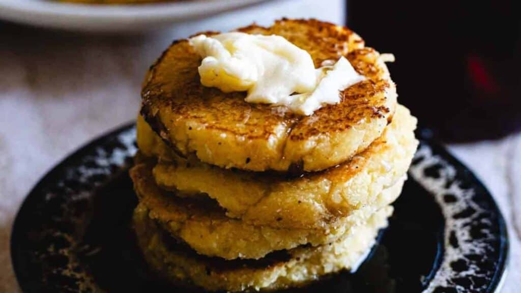 A stack of four golden-brown cornmeal cakes topped with a dollop of butter, served on a dark plate.