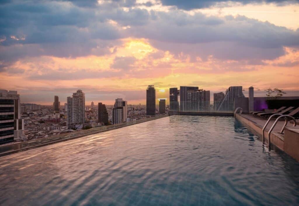 Rooftop infinity pool at a Bangkok hotel overlooking the city skyline at sunset, with tall buildings and a partly cloudy sky in the background.