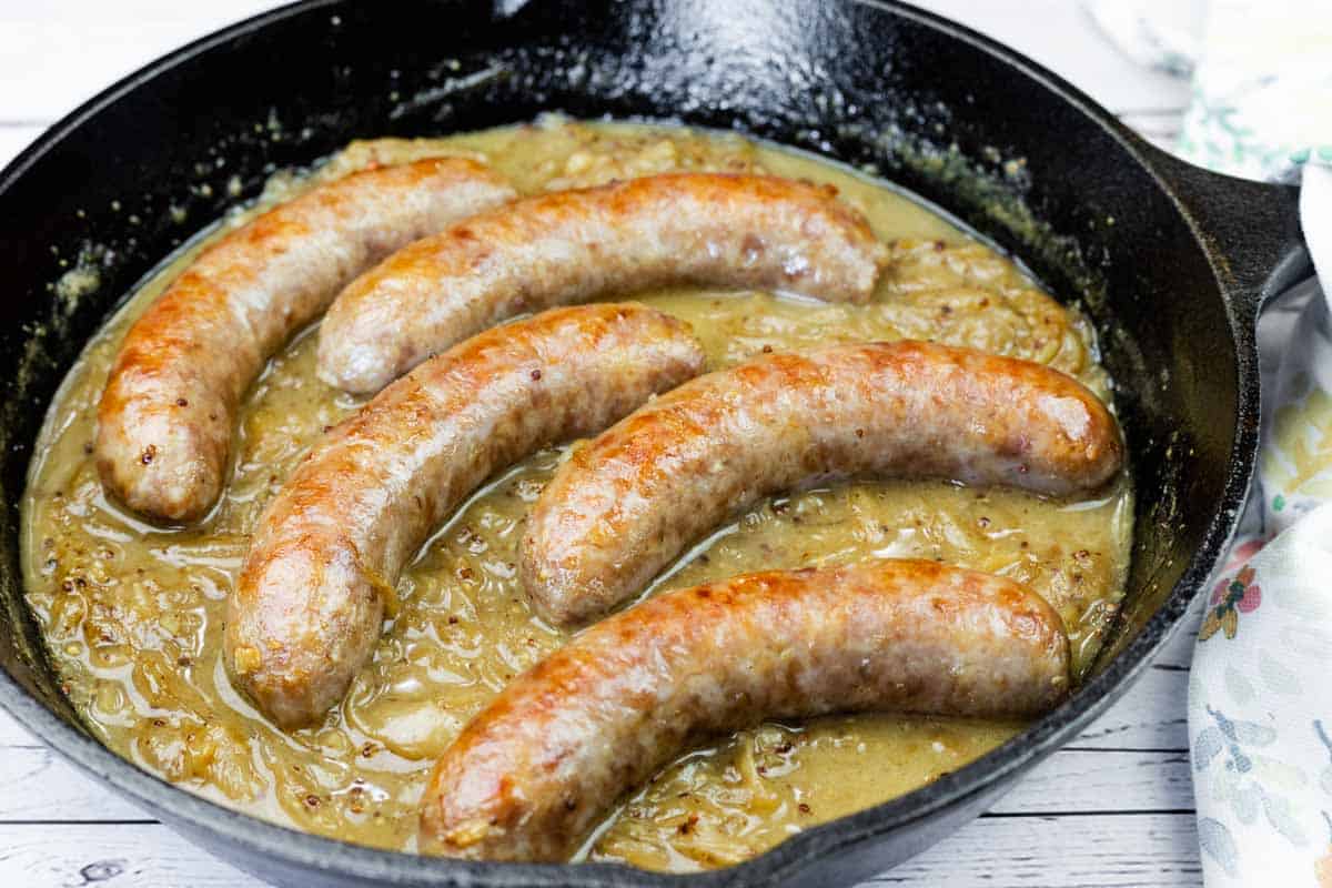 Five browned sausages simmer in onion gravy inside a black cast iron skillet on a light wooden surface.