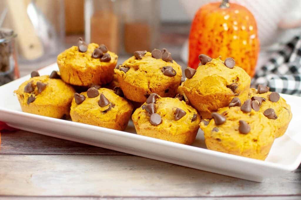 A rectangular white plate holds several pumpkin muffins topped with chocolate chips. A small decorative pumpkin is visible in the background.