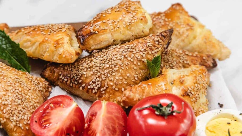 Golden, sesame-topped triangular pastries are stacked on a tray, with fresh tomato halves and a sliced hard-boiled egg beside them.