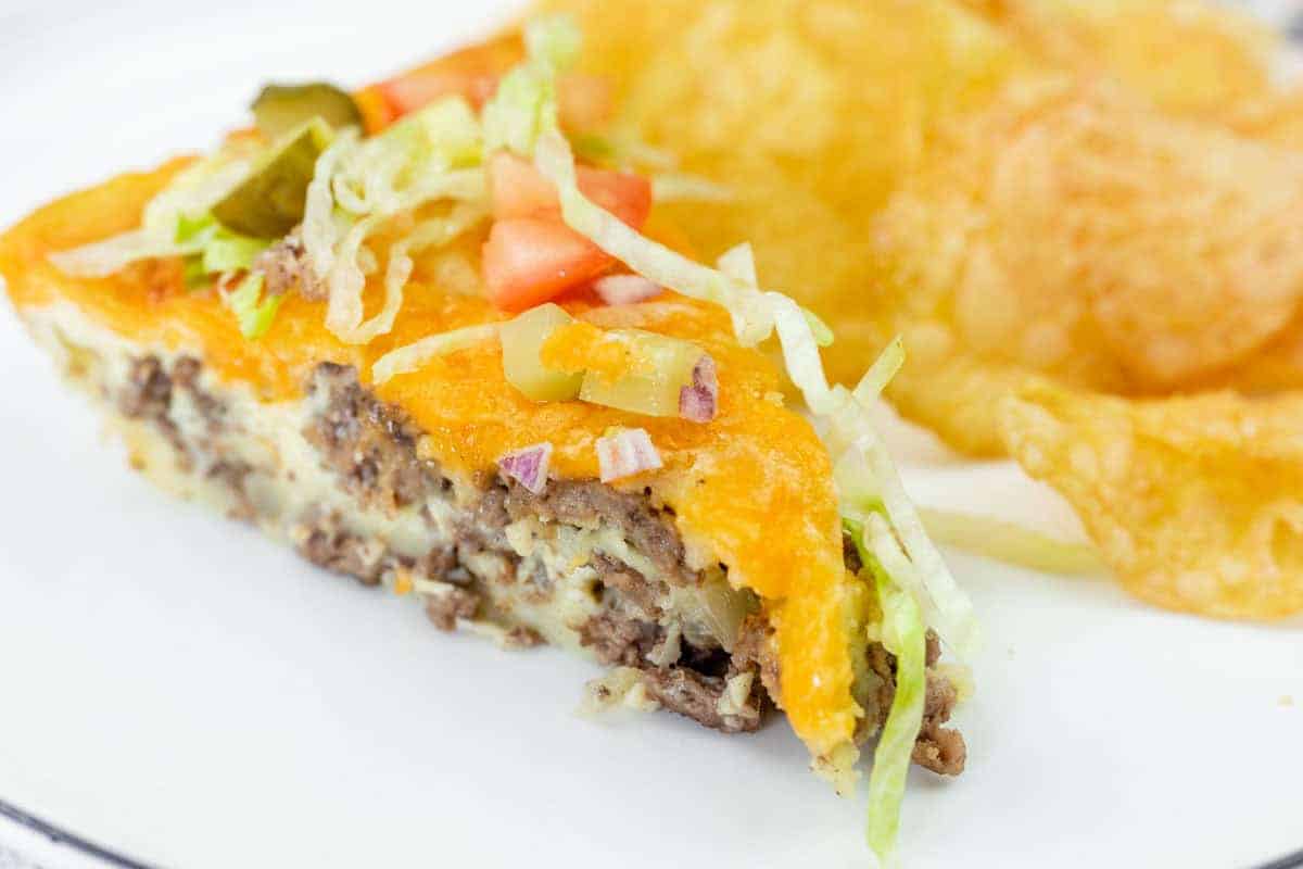A slice of cheeseburger pie topped with shredded lettuce, diced tomato, onions, and pickles, served with potato chips on a white plate.