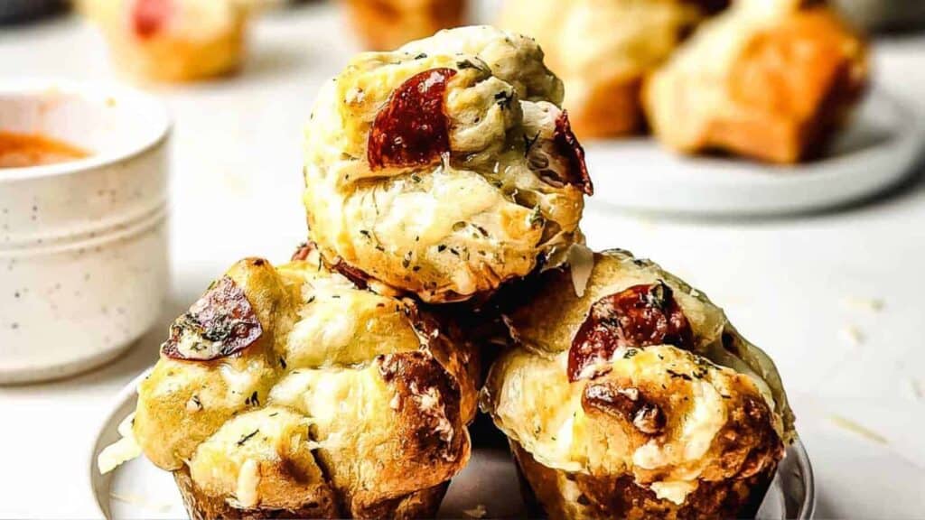 Three savory muffins topped with melted cheese and pepperoni slices are stacked on a plate, with a cup of dipping sauce and more muffins in the background.