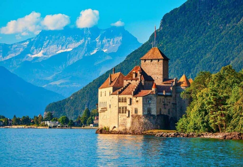 A stone castle with red roofs sits on the edge of a lake, surrounded by green hills and distant mountains under a blue sky with clouds.
