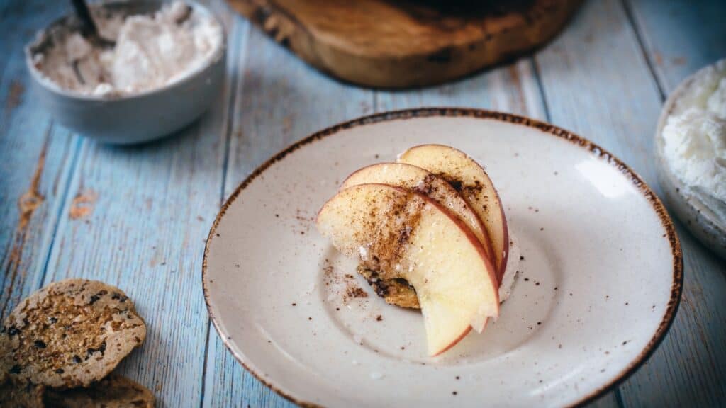Sliced apples arranged on a small plate, sprinkled with cinnamon, with crackers and spread in bowls nearby on a rustic wooden table.