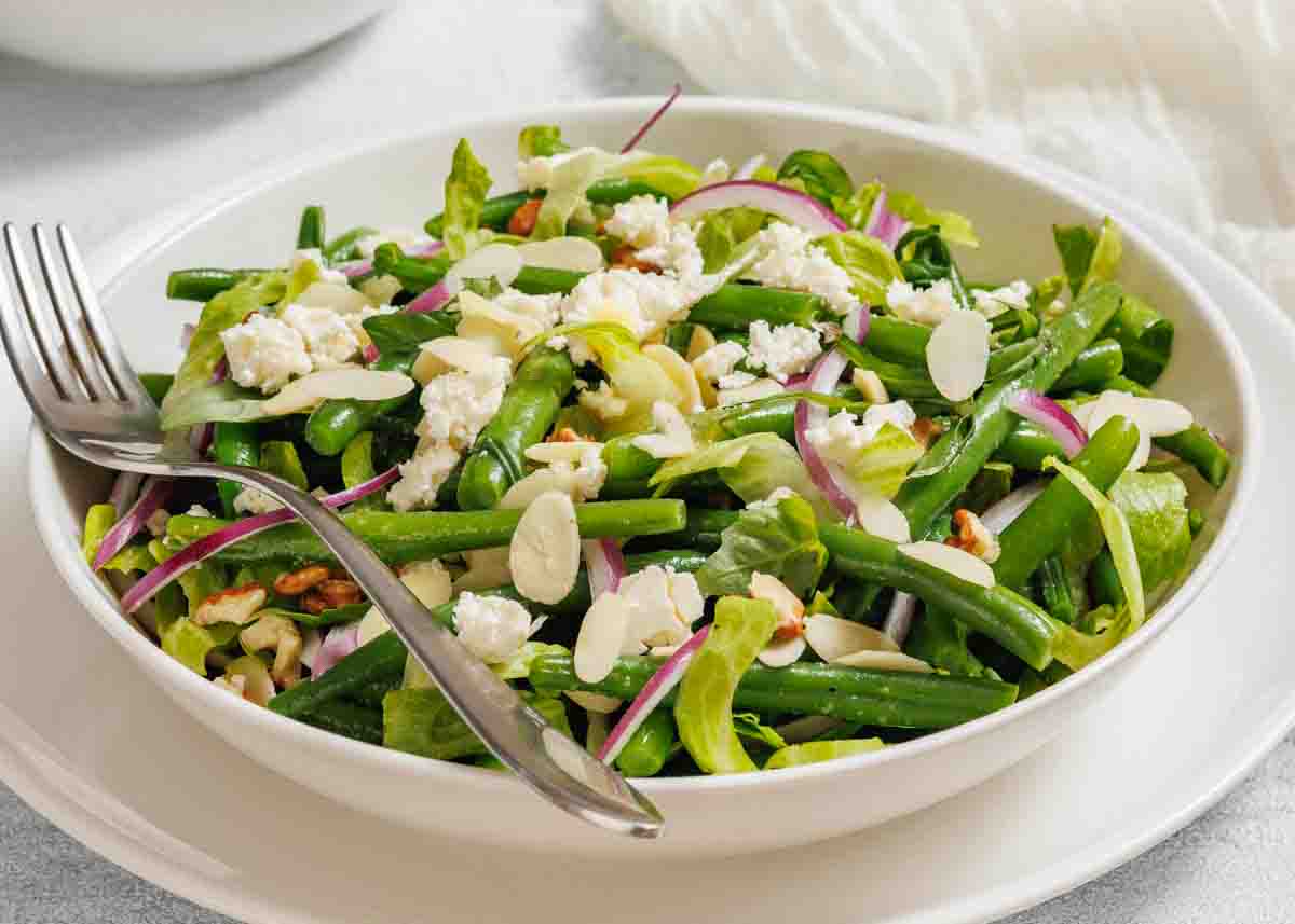 A bowl of green bean salad with feta cheese, sliced almonds, red onions, and lettuce, served with a fork and spoon.