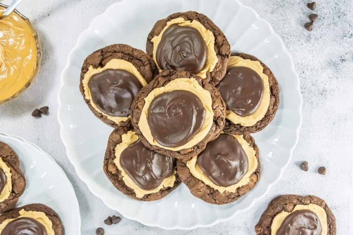 A white plate holds several chocolate cookies topped with a layer of peanut butter and a swirl of chocolate ganache.