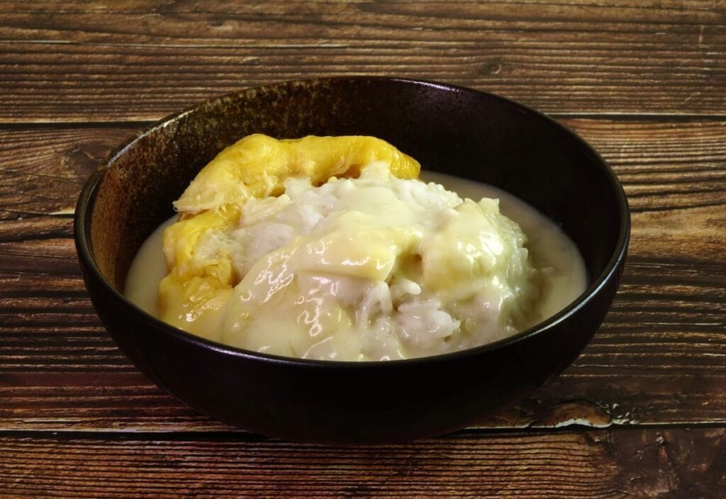 A black bowl filled with sticky rice and sliced durian topped with coconut cream sauce, placed on a wooden surface.