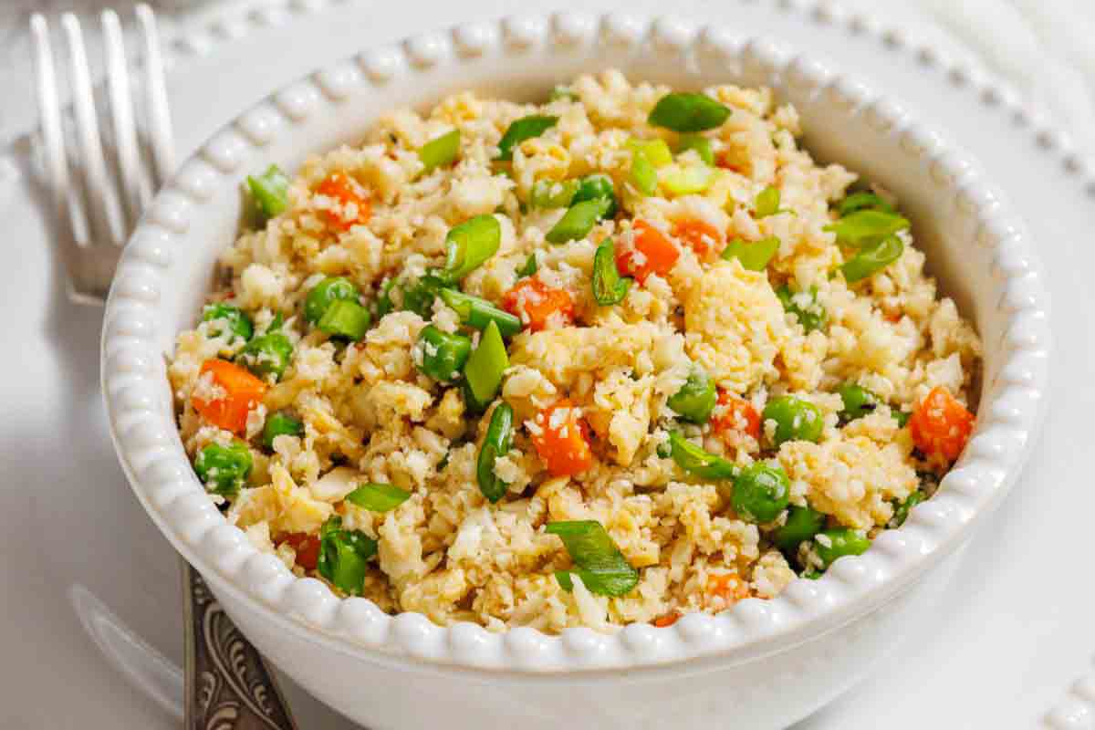 A white bowl contains cauliflower rice mixed with chopped carrots, peas, and green onions. A fork is placed beside the bowl on a white plate.