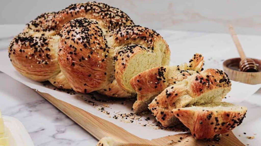 A braided loaf of bread topped with sesame and black seeds, partially sliced on a wooden board, with honey and butter in the background.