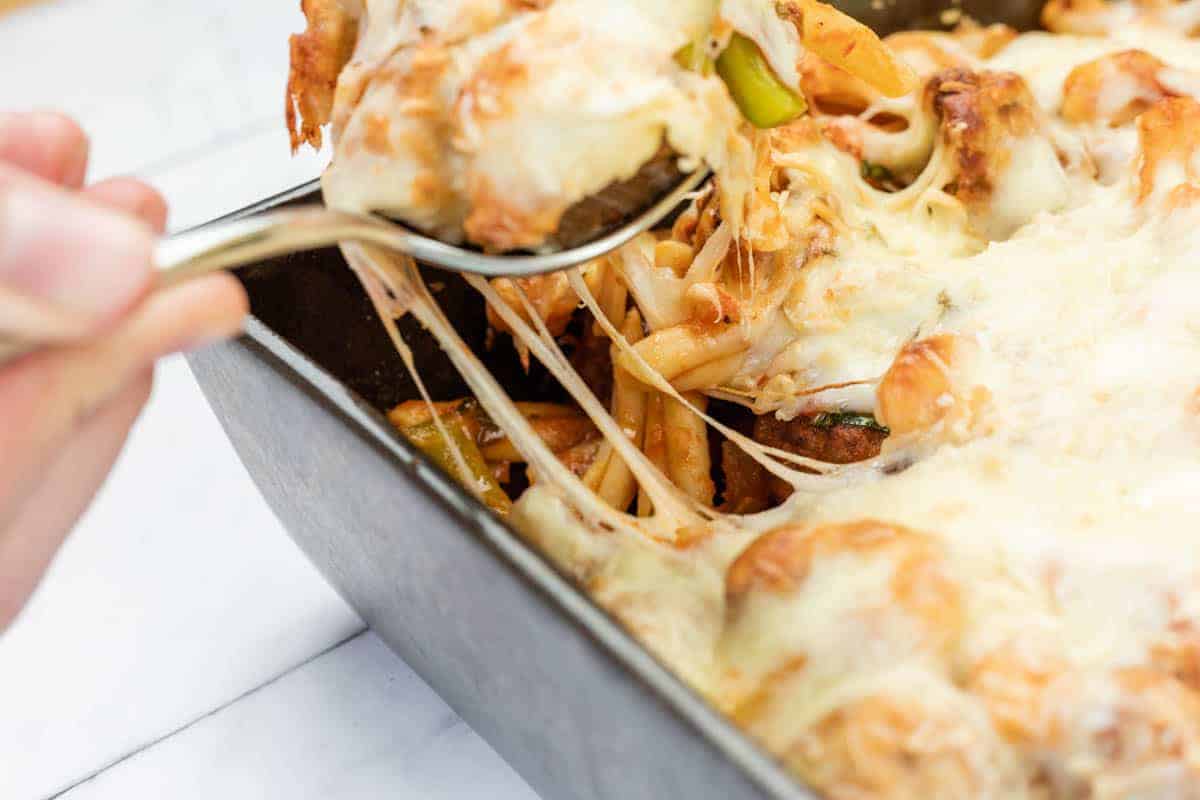 A hand uses a spoon to serve Easy Cheesy Meatball Casserole from a rectangular baking dish, with melted cheese stretching from the dish.