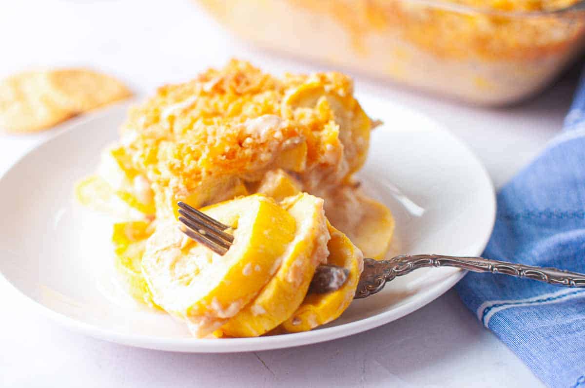 A serving of yellow squash casserole topped with a crumbly crust sits on a white plate with a fork, next to a blue cloth napkin.