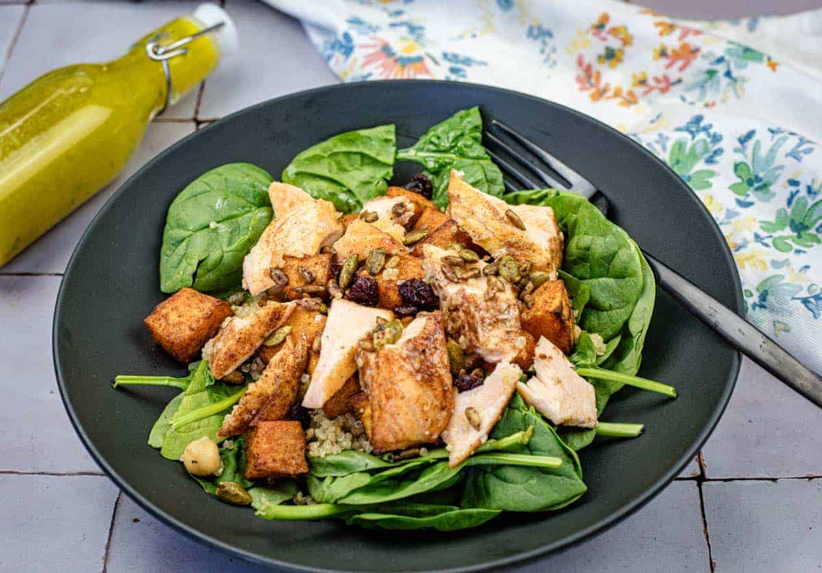 A black plate with spinach leaves, grilled chicken pieces, roasted sweet potatoes, nuts, seeds, and quinoa. A fork and a bottle of yellow dressing are nearby.