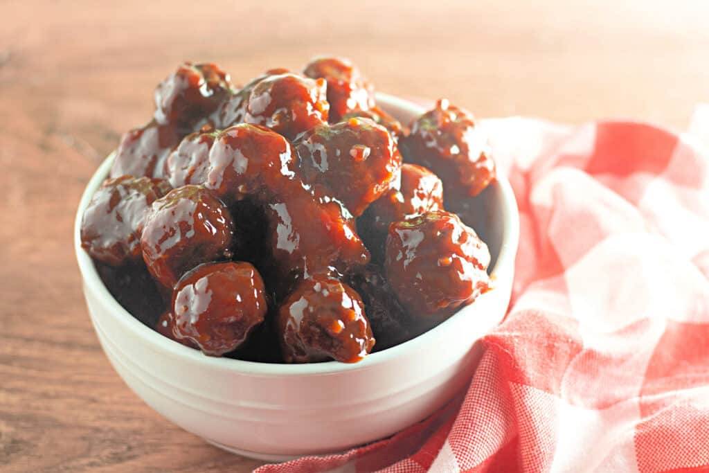 A white bowl filled with glazed meatballs sits on a wooden surface next to a red and white checkered cloth.