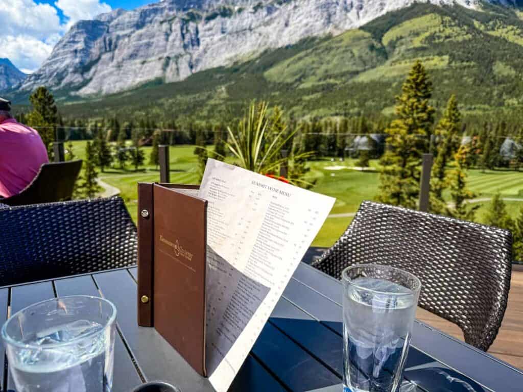 A restaurant menu and two glasses of water sit on an outdoor table overlooking a golf course and mountains under a partly cloudy sky, setting the scene for a five star meal.