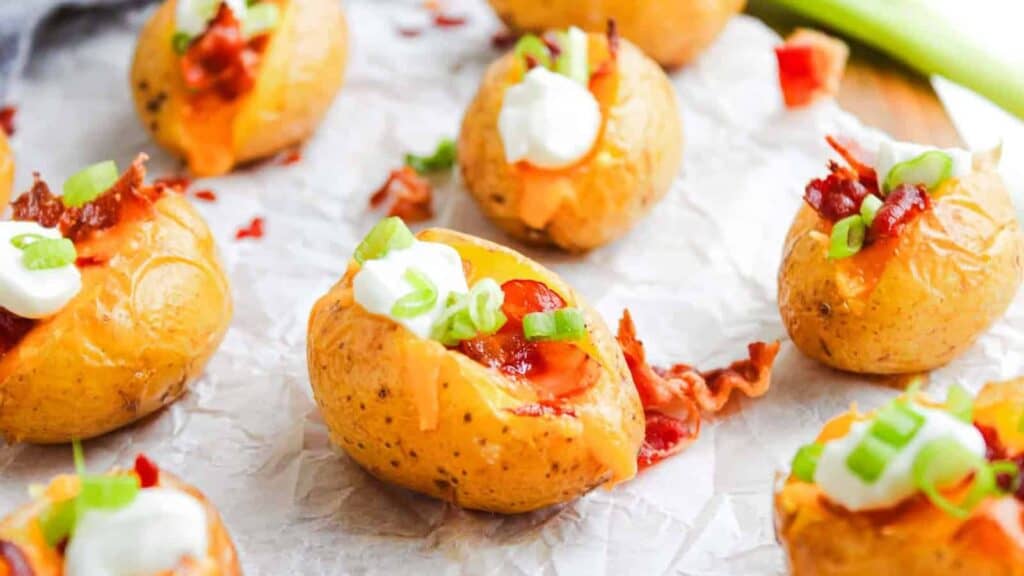 Mini baked potatoes topped with melted cheese, bacon bits, sour cream, and chopped green onions, arranged on parchment paper.