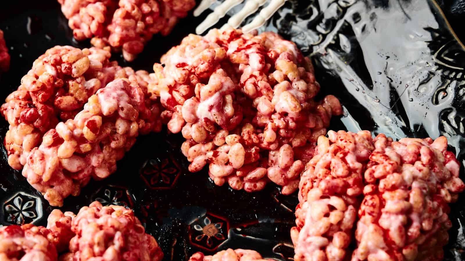 Clusters of pink and red puffed rice cereal treats are arranged on a dark, patterned plate next to a white skeleton hand decoration.