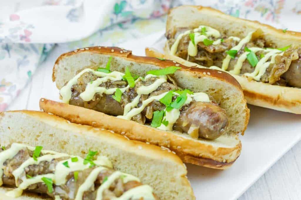 Three sausage sandwiches with grilled onions, parsley, and a drizzle of mustard on toasted buns, served on a white tray.