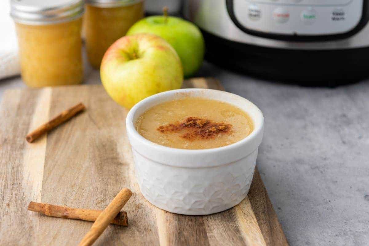 A white ramekin filled with applesauce topped with cinnamon sits on a wooden board next to cinnamon sticks, apples, and an Instant Pot.