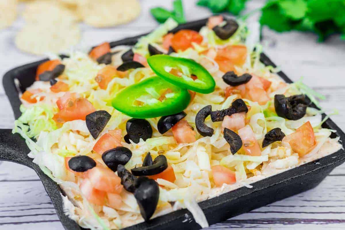 A skillet of Taco dip filled with shredded lettuce, diced tomatoes, sliced black olives, and jalape&ntilde;o slices, served on a light wooden surface.