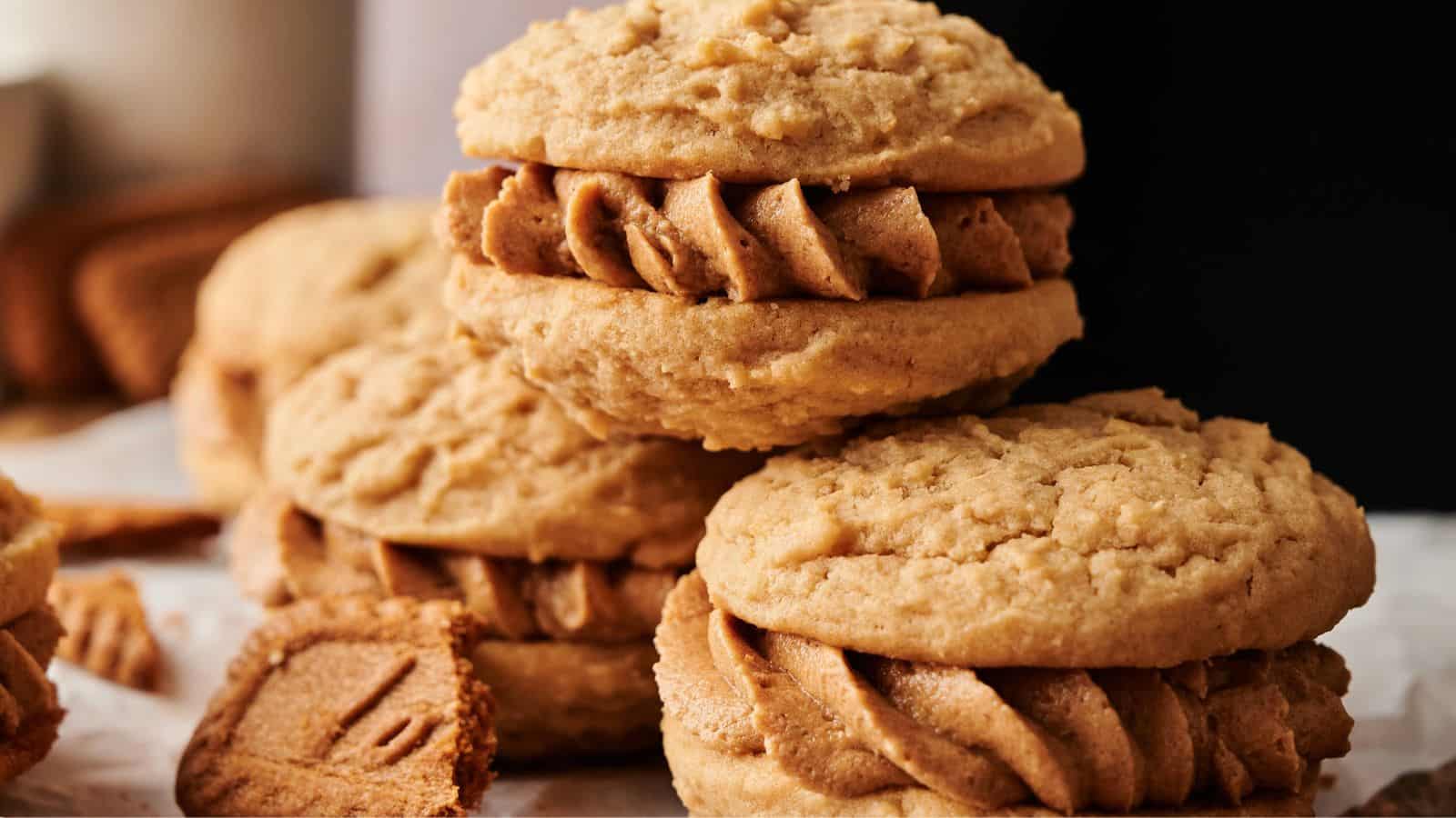 Three sandwich cookies filled with light brown frosting are stacked on a surface, with broken cookie pieces scattered nearby.