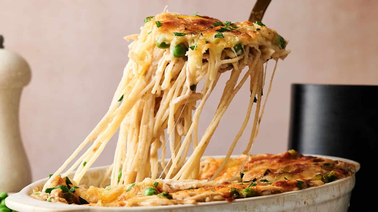 A serving of baked spaghetti with melted cheese, peas, and herbs is being lifted from a casserole dish.